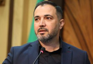 یک فرد در حال سخنرانی پشت میکروفون با کت رسمی تیرهرنگ، در محیطی رسمی با پسزمینه چوبی و پرچم.