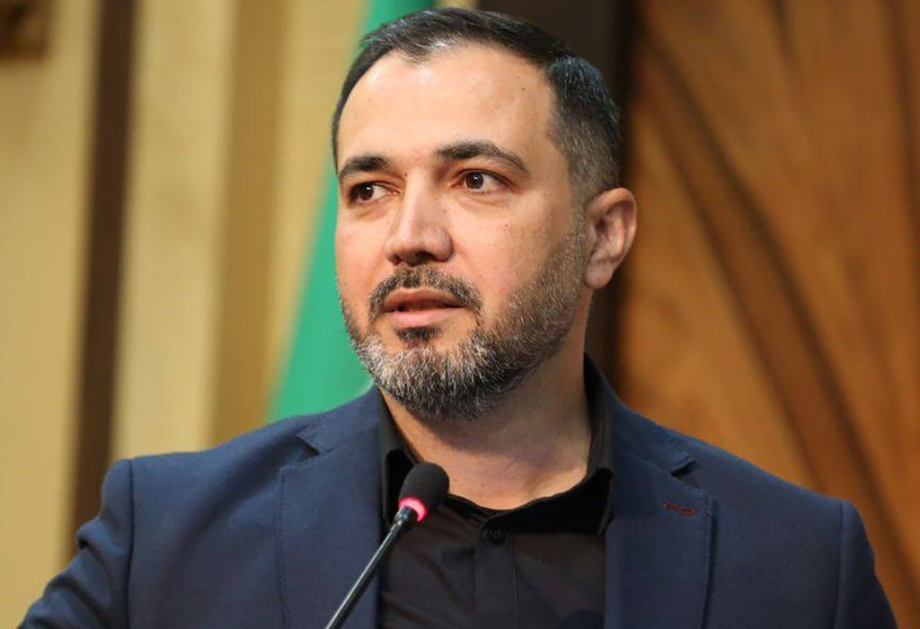 یک فرد در حال سخنرانی پشت میکروفون با کت رسمی تیرهرنگ، در محیطی رسمی با پسزمینه چوبی و پرچم.